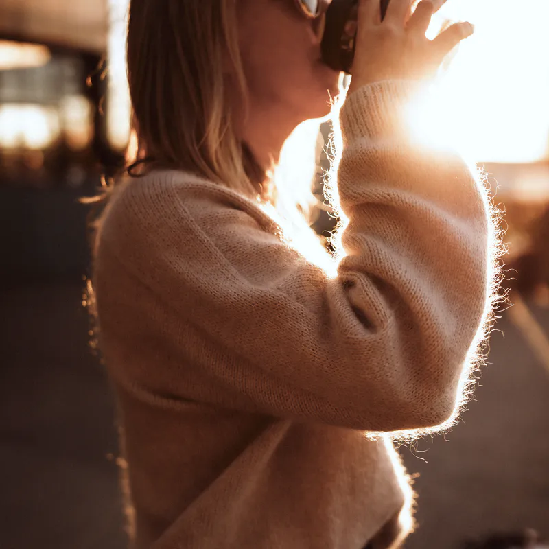 The sun peeks past the raised arm of a woman drinking a coffee late in the day which is always a silly idea. The seamless sleeve detail of her hand knit is the focus