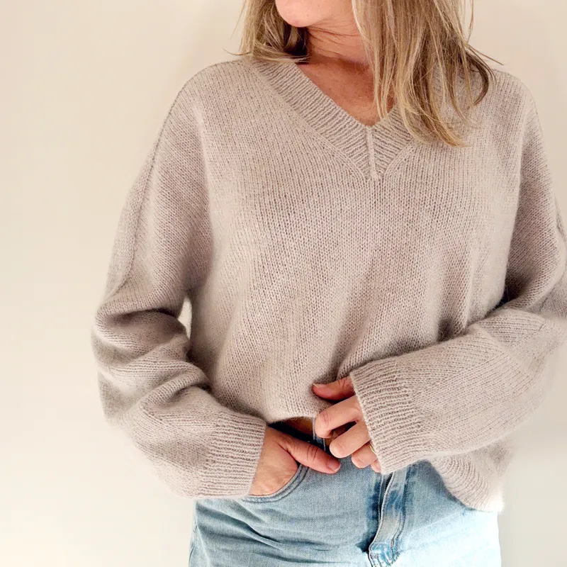 A woman wearing a beige, hand knit sweater. It is a V-neck design with a saddle shoulder that runs seamlessly to the cuff