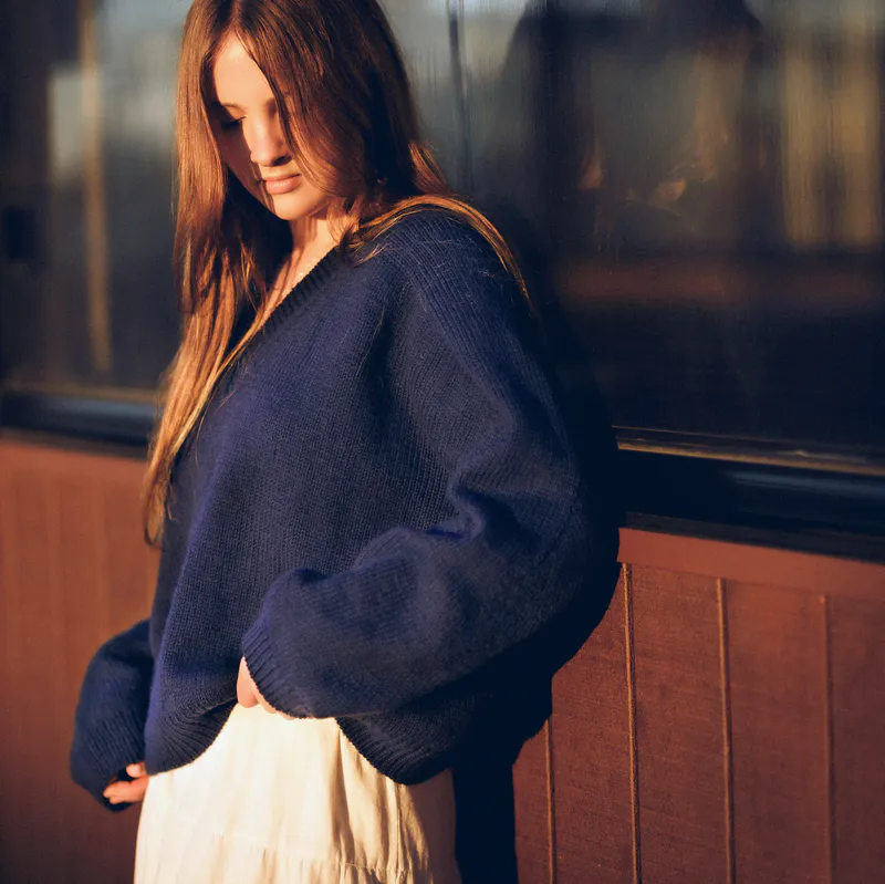 A woman wearing the V-neck Ridgeline Sweater in the setting sun. She's looking at the ground and a brown wall with windows is behind her.