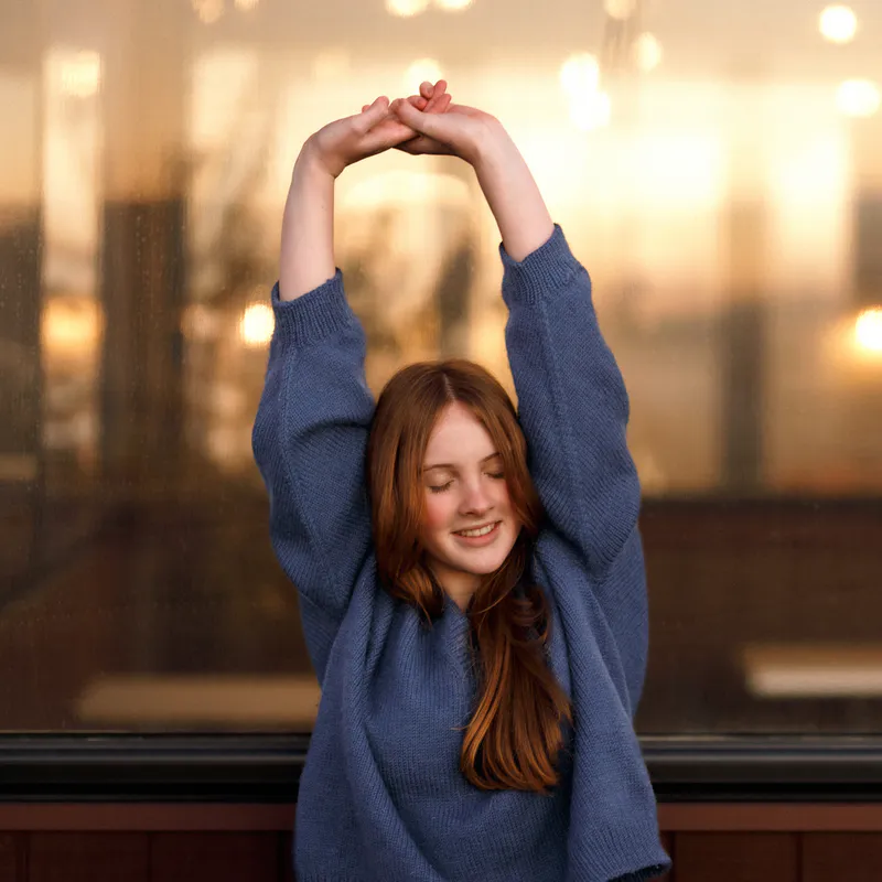 A woman with her arms raised above her head. She wears a blue hand knit sweater and the under-sleeve details are showing as a ridge of decrease stitches