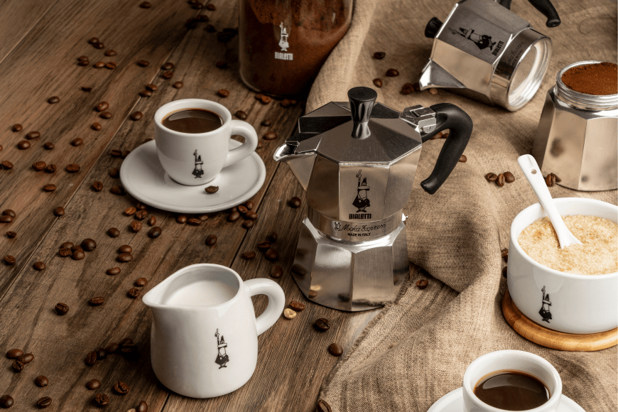 There are two Bialetti moka pot stovetop coffee makers in the picture, one is separated in half with ground coffee showing in the bottom basket. There is a small white milk jug with the Bialetti logo on the front and a matching sugar bowl. There are coffee beans scattered around. Set on a lightly coloured wooden table