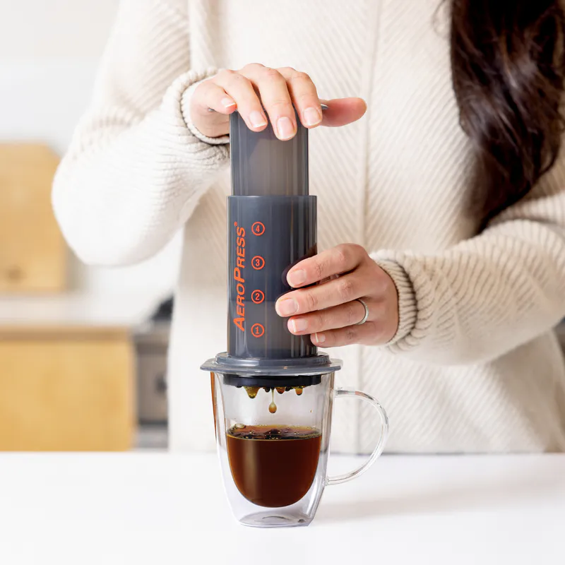 Woman in white sweater making coffee with AeroPress in kitchen