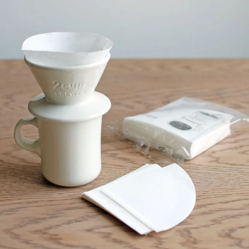 Kinto Coffee Filter inside a 2 cup white ceramic brewer sitting on top of a white ceramic mug. There is a spare packet of filters in the background