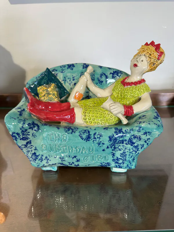 A Ceramic sculpture of a birthday girl reclining on the sofa with a bottle of bubbles by Mandy Olson, available at The Little Gallery Tairua