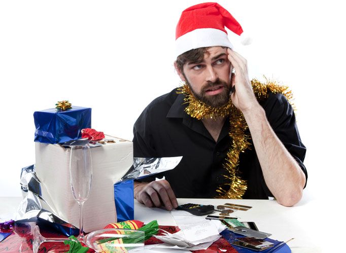 Stressed-looking man wearing a Santa hat and tinsel around his neck sits at a table laden with Christmas gifts
