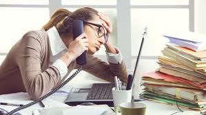 Woman sitting at desk cluttered with files has phone to her ear and looks stressed