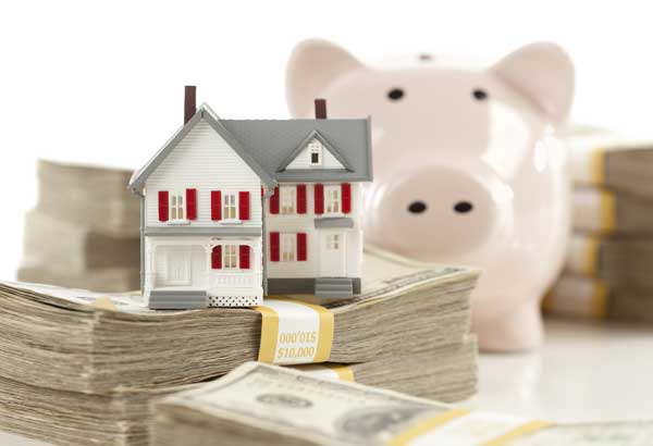 Model of a house sitting on bundles of cash with a piggy bank in the background